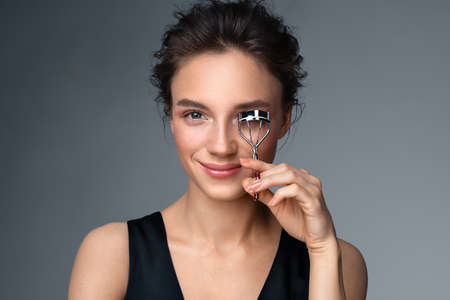 Beautiful woman using eyelash curler on long eyelashes. Photo of woman with perfect makeup on gray background. Beauty conceptの写真素材