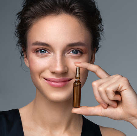 Woman holds ampoule with serum for hair or skin care. Photo of attractive woman with perfect makeup on gray background. Beauty conceptの写真素材