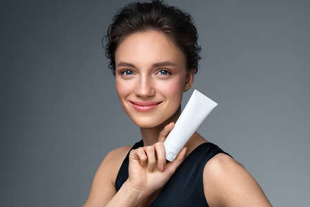Woman holds tube with cosmetic cream. Photo of attractive woman with perfect makeup on gray background. Beauty conceptの写真素材