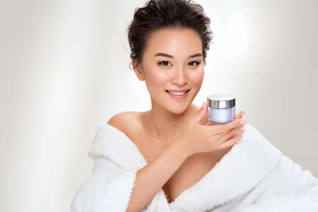 Woman holds jar with cosmetic cream. Photo of asian woman after shower on white background. Beauty and skin care conceptの写真素材