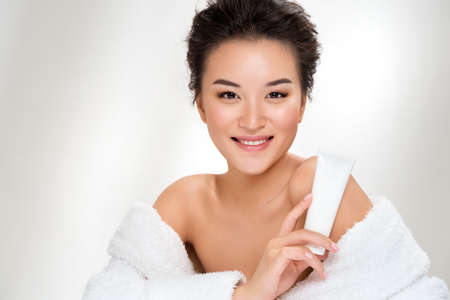 Woman holds tube with cosmetic cream. Photo of asian woman after shower on white background. Beauty and skin care conceptの写真素材