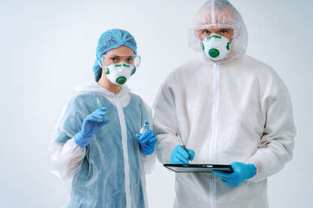 Doctor holds clipboard and nurse holds an injection syringe and vaccine. Healthcare workers in protective suits and medical masksの写真素材