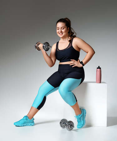Sporty girl workout with dumbbells. Photo of model with curvy figure in fashionable sportswear on gray background. Sports motivation and healthy lifestyleの写真素材
