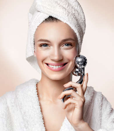 Attractive woman massaging her cheek with massage Y-shaped ball roller. Photo of woman with perfect skin on beige background. Beauty and skin care conceptの写真素材