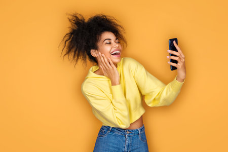 Young girl holds smartphone and takes selfie photo, smiles at camera. Photo of african american girl on yellow backgroundの写真素材