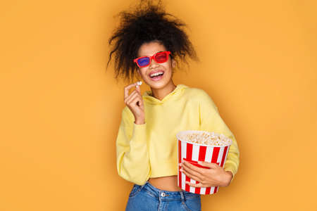 Young girl watching tv with popcorn box. Photo of african american girl on yellow backgroundの写真素材