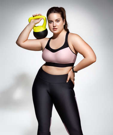 Strong woman workout with kettlebell. Photo of model with curvy figure in fashionable sportswear on gray background. Sports motivation and healthy lifestyleの写真素材