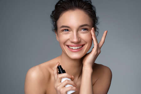 Beautiful woman holds bottle with cosmetic cream and touches her perfect skin on gray background. Beauty and skin care conceptの写真素材