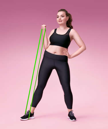 Strong girl working with resistance band. Photo of model with curvy figure in fashionable sportswear on pink background. Sports motivation and healthy lifestyleの写真素材