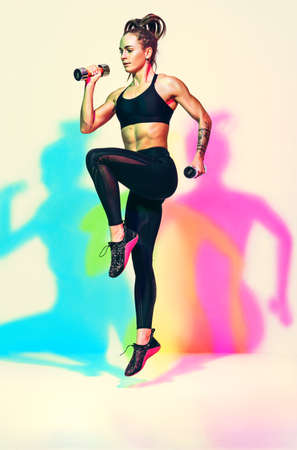 Sporty girl jumping with dumbbells. Photo of muscular woman in black sportswear on white background with effect of rgb colors shadows. dynamic movement. Sports motivation and healthy lifestyleの写真素材