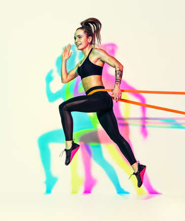 Sporty woman doing jumping exercise with resistance band. Photo of muscular woman in black sportswear on white background with effect of rgb colors shadows. dynamic movement. side view. Sport and healthy lifestyleの写真素材