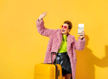 Happy woman is ready to travel. Photo of elderly woman wears pink glasses, dressed in fashionable outfit, with tickets and suitcase on yellow backgroundの写真素材