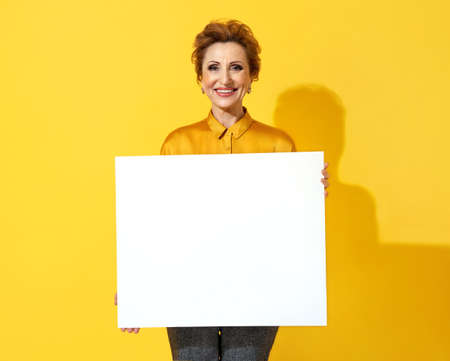 Smiling woman with white empty board. Photo of attractive elderly woman in yellow shirt on yellow backgroundの写真素材