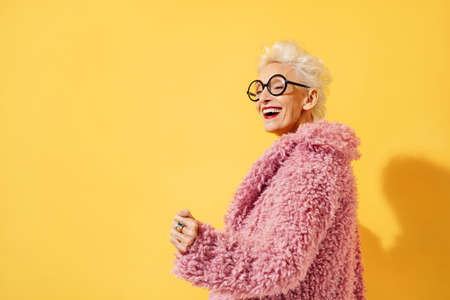 Happy and playful mature woman dancing, smiling and having fun. Photo of elderly woman over 70 years old in stylish outfit on yellow backgroundの写真素材