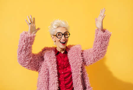 Happy and playful mature woman dancing, smiling and having fun. Photo of elderly woman over 70 years old in stylish outfit on yellow backgroundの写真素材