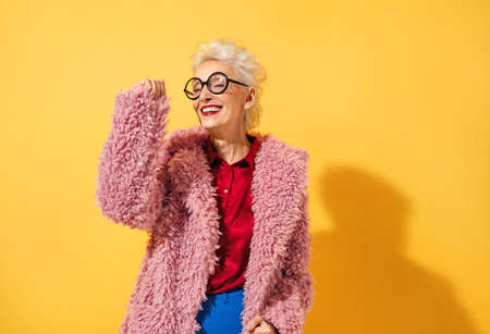 Happy and playful mature woman dancing, smiling and having fun. Photo of elderly woman over 70 years old in stylish outfit on yellow backgroundの写真素材