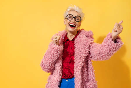 Happy and playful mature woman dancing, smiling and having fun. Photo of elderly woman over 70 years old in stylish outfit on yellow backgroundの写真素材