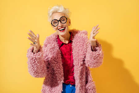 Happy and playful mature woman dancing, smiling and having fun. Photo of elderly woman over 70 years old in stylish outfit on yellow backgroundの写真素材