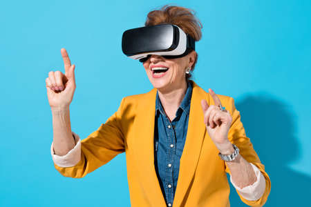 Smiling woman interacting with invisible screen during work in virtual space. Photo of mature woman with modern VR goggles in yellow blazer touching air on blue backgroundの写真素材