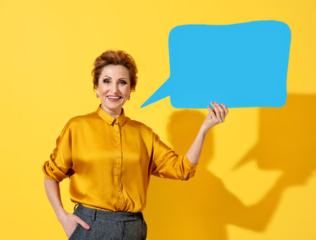 Happy mature woman holding blue speech bubble banner. Photo of kind attractive woman in yellow shirt on yellow backgroundの写真素材
