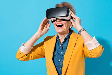 Happy mature woman with modern VR goggles. Photo of business woman in yellow blazer on blue backgroundの写真素材
