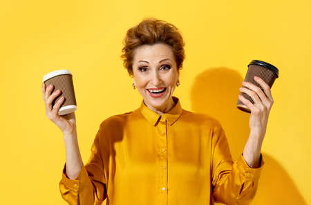 tea or coffee? Joyful woman holds in both hands paper cups to choose from. Photo of elderly woman in yellow shirt on yellow background. Emotions and pleasant feelings concept.の写真素材