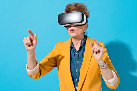 Business woman interacting with invisible screen during work in virtual space. Photo of mature woman with modern VR goggles in yellow blazer touching air on blue backgroundの写真素材