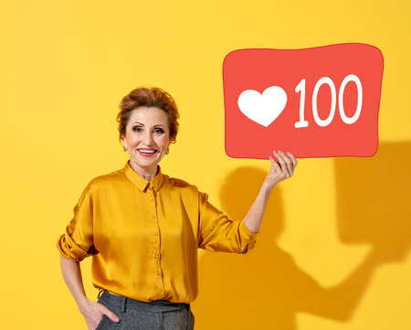 Happy mature woman holding red board with 100 likes. Photo of kind attractive woman in yellow shirt...