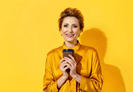 Attractive mature woman holding hot takeaway coffee. Photo of smiling woman in yellow shirt on yellow background. Emotions and pleasant feelings concept.の写真素材