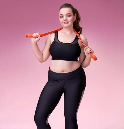Sporty girl with resistance band. Photo of pretty model with curvy figure in black sportswear on pink background. Sports motivation and healthy lifestyleの写真素材