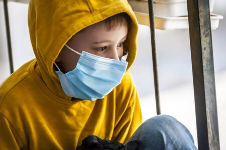 Cute Caucasian school age boy in a surgeon protection mask sitting by the window with bars. Coronavirus corona virus quarantine, no school, not allowed to play outside, forced to sit at home.の写真素材