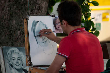 A street artist sits back on a chair in a red t-shirt in summer. And on the street and draws a portrait of a girl from a photoのeditorial素材