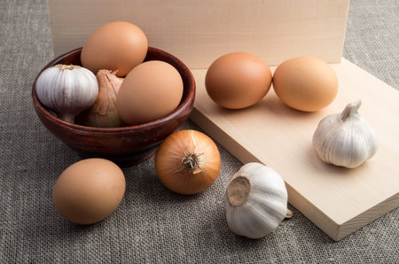 Still-life on the gray sacking - raw eggs, onions and garlic next to an old wooden bowl on a background of textileの写真素材