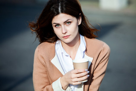 Young stylish woman drinking tea in a city street. Hipster european girl with latte paper glass in street.の写真素材