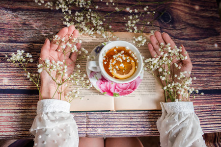 A cup of tea in the hands of a girl with a book on a wooden backgroundの写真素材