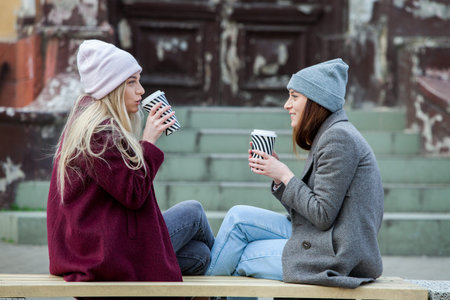 Two young women sitting on a bench and drinking coffee in the city