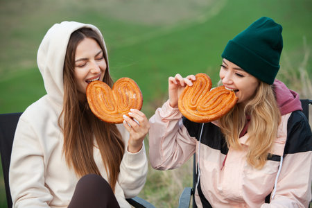 Two beautiful girls in warm clothes and hats eat buns on the nature