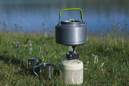 Camping stove with a kettle and two cups on the grass near the lake
