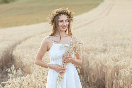 Portrait of a beautiful young woman in white dress and wreath of wheat ears in the fieldの写真素材