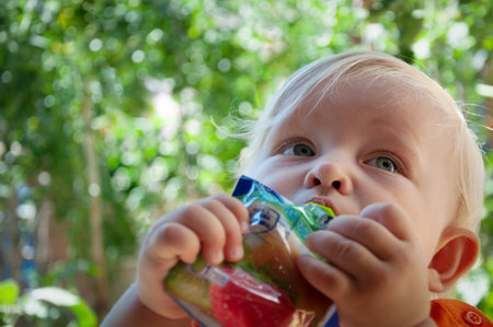 little blonde girl eats fruit puree in the park in summer, healthy baby foodの写真素材