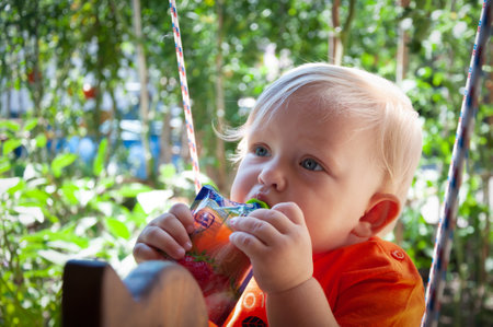 little blonde girl eats fruit puree in the park in summer, healthy baby foodの写真素材