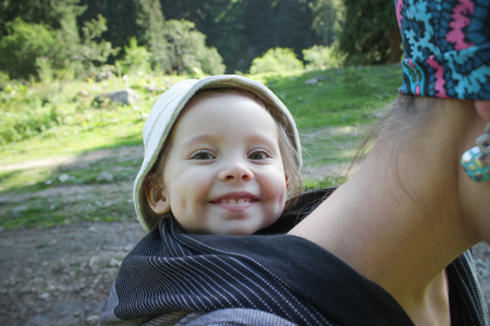 Mother carrying a two year old smiling baby in a black woven wrap. Tracking, outdoorの写真素材