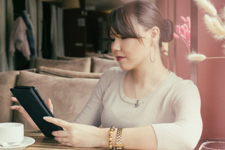 An attractive young asian woman sitting in a cafe is holding a tablet in her hand