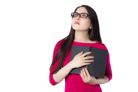 Smart young student woman in red shirt and glasses pressing black book to her heart, looking above on white background, knowledge woman concept ideaの写真素材