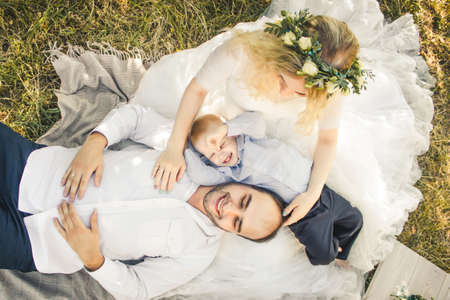 Little blond haired toddler boy, groom lying on knees of bride in wedding dress. Outdoor wedding scene in sunny summer dress.の写真素材
