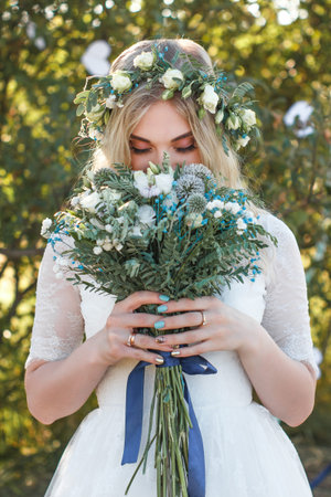 Beautiful blonde young bride portrait with flower bouquet and wreath on her head in white wedding...