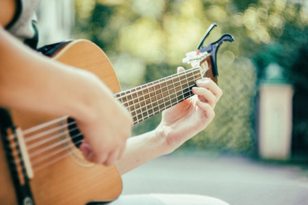 Close up guitar neck with capo in the park. Young man sitting on the bench in the park playing the guitar. Young attractive man enjoys live music in last sunny days autumn holiday. Retro lens used.の写真素材