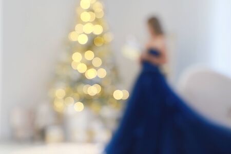 Blur light celebration on christmas tree with white wall background. Woman in elegant blue dress holds a Christmas present in her hands. Defocus holiday background.の写真素材