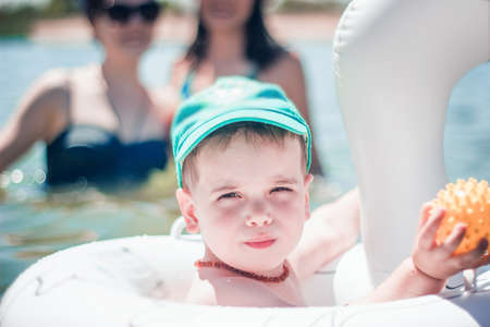 A cute little boy in a blue baseball hat swimming in the sea in an inflatable ring.の写真素材