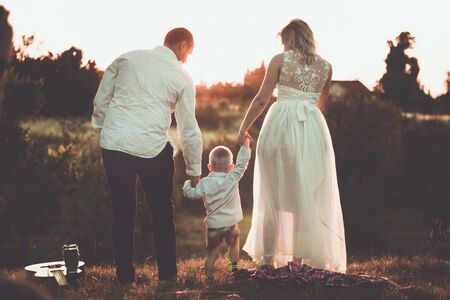 Little family romantic picnic scene outdoor in sunset. Parents holding hands of little toddler boy between them.の写真素材
